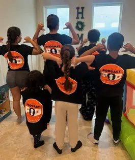 Group of kids and adults proudly wearing Podcast Pod Nashville t-shirts, facing away to show large orange back logo, standing together in a home